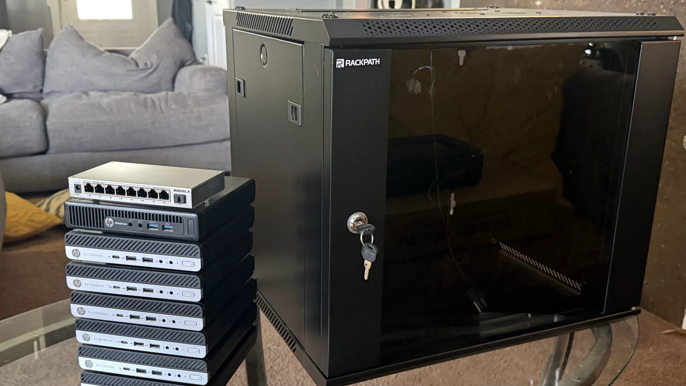 A stack of seven mini PCs topped by a network switch, next to a closed black wall cabinet